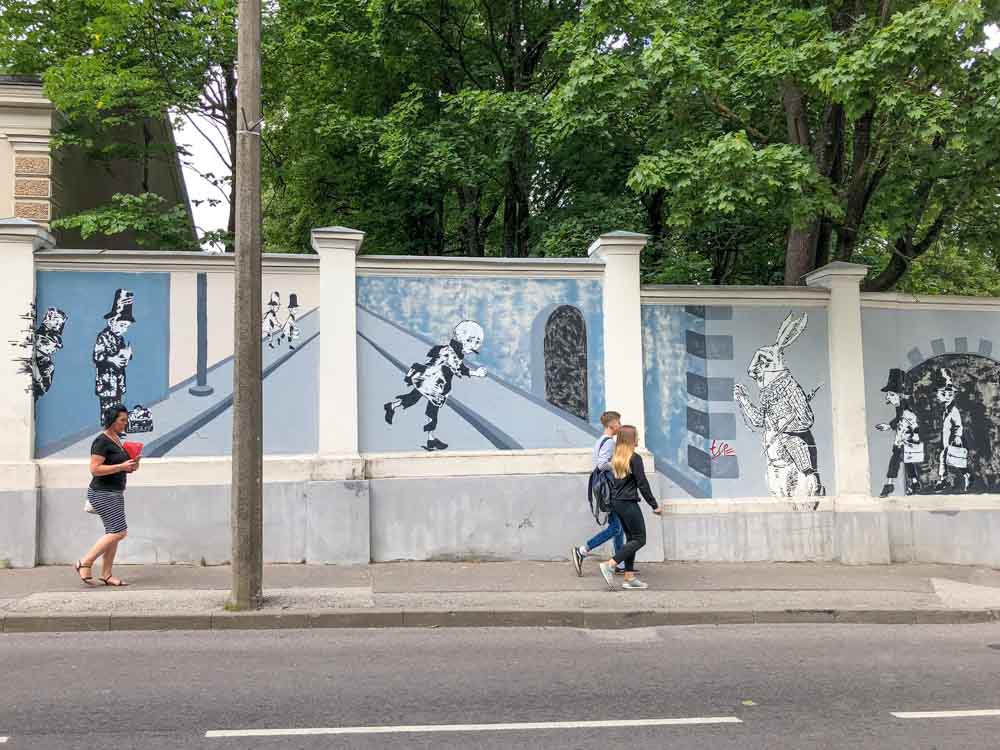 Tartu children's story mural by Hare. blue street art with people walking on sidewalk.